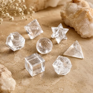 May include: A collection of seven clear quartz crystal geometric shapes including a cube, a sphere, a pyramid, a star, and various polyhedrons. Each piece is transparent with natural internal inclusions, arranged on a textured tan surface.