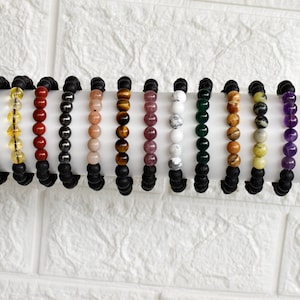 Puede incluir: Un conjunto de 15 pulseras con cuentas de piedras preciosas de varios colores y cuentas de lava negra. Las pulseras están dispuestas en fila sobre una superficie blanca.