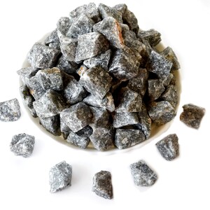 May include: A white bowl filled with gray, irregularly shaped rock salt crystals. Several smaller crystals are scattered around the bowl on a white surface.