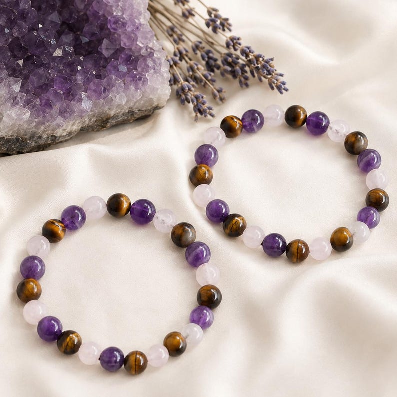 May include: Two circular beaded bracelets featuring a repeating pattern of polished spherical beads in shades of purple amethyst, brown tiger eye, and pale pink rose quartz. The bracelets are arranged on a soft, off-white fabric surface.