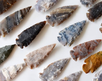 May include: A collection of various stone arrowheads, showcasing a range of colors and patterns. The arrowheads are in shades of gray, brown, and tan, with some displaying unique veining and textures. The collection is arranged on a white surface.
