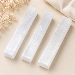 May include: Three rectangular selenite wands with a white, fibrous, and translucent appearance. Each wand features a smooth, elongated shape with a distinct linear texture running along the length of the stone, reflecting light across the surface.