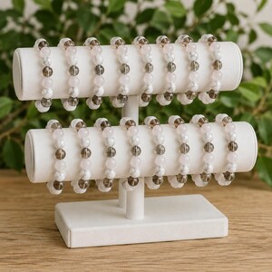 Puede incluir: Un expositor de joyas blanco de dos niveles con diecis&eacute;is pulseras de cuentas. Cada pulsera presenta un patr&oacute;n repetitivo de cuentas redondas pulidas en gris humo transl&uacute;cido, blanco opaco con vetas grises y cuarzo rosa p&aacute;lido.