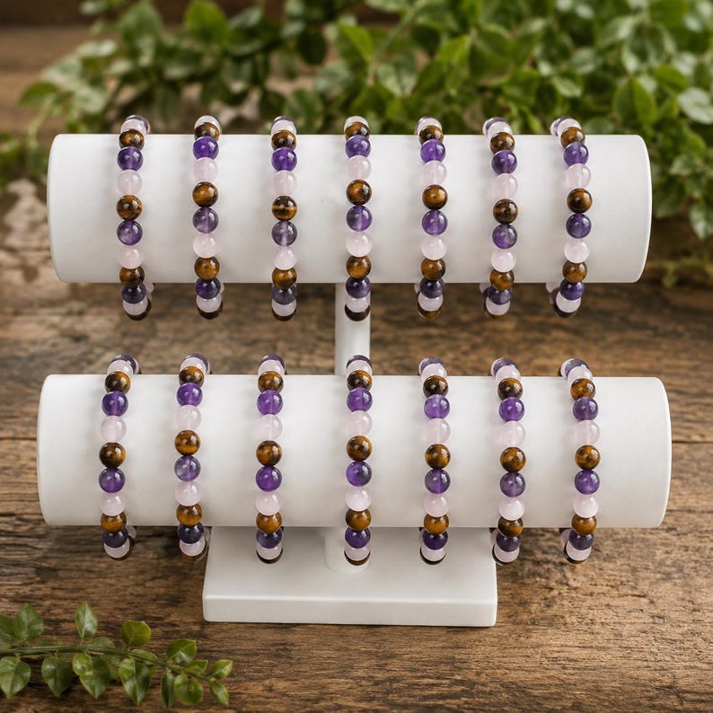 May include: A collection of fourteen beaded bracelets displayed on a two-tiered white stand. Each bracelet features a repeating pattern of round polished beads in shades of purple, light pink, and brown tiger eye, strung on an elastic cord.