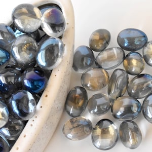 May include: A bowl filled with iridescent glass pebbles in shades of blue and gray. The pebbles are scattered around the bowl and on a white surface.