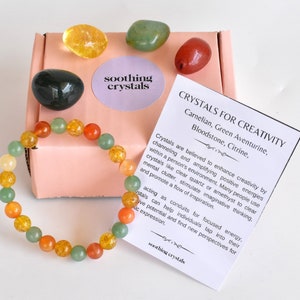May include: A pink box with a variety of crystals, including carnelian, green aventurine, bloodstone, and citrine. The box also includes a bracelet made with the same crystals. The text on the card reads "Crystals for Creativity".