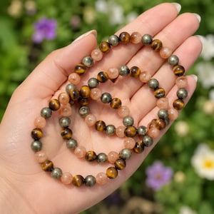 Pode incluir: Duas pulseiras de contas com pedras redondas polidas alternadas em tons de olho de tigre castanho-dourado, pirite metálica e pedra do sol translúcida cor de pêssego. As contas estão enfiadas num fio elástico para formar joalharia.
