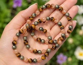May include: Two beaded bracelets featuring alternating round polished stones in shades of golden-brown tiger's eye, metallic pyrite, and translucent peach-colored sunstone. The beads are strung on an elastic cord to form circular wrist jewelry.