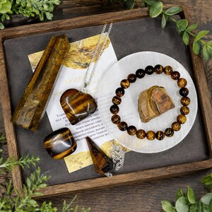 May include: A collection of tiger's eye items including a tall polished crystal tower, a heart-shaped pendant on a silver chain, a smooth tumbled stone, a beaded bracelet, a pointed pendant, and a raw stone piece resting on a white circular base.