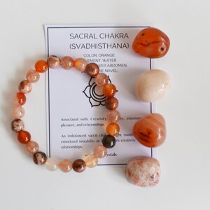 May include: Orange carnelian crystal bracelet and tumbled stones. The card reads "Sacral Chakra (Svadhisthana)" and lists the colour, element, and location of the chakra. Associated with creativity, pleasure, and relationships.
