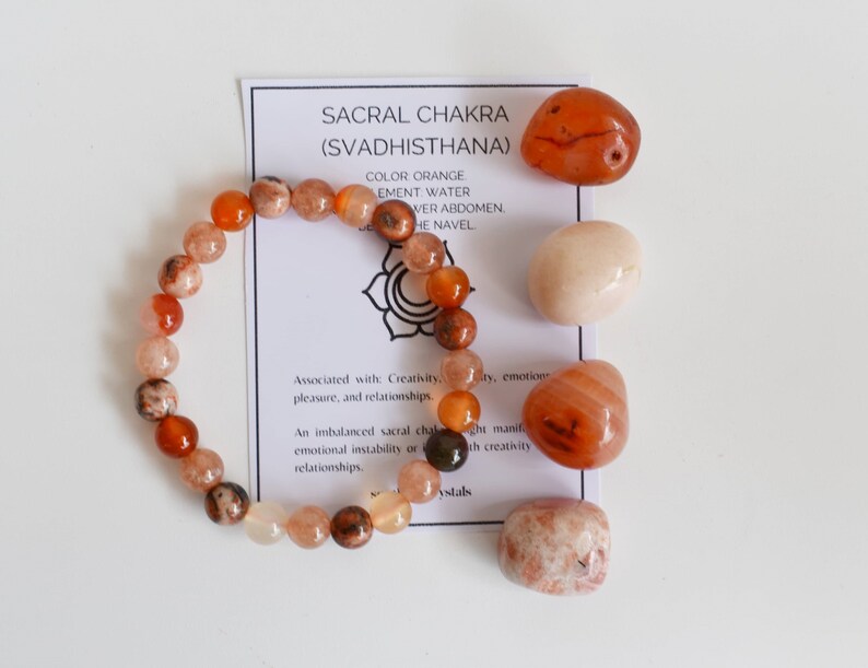 May include: Orange carnelian crystal bracelet and tumbled stones. The card reads "Sacral Chakra (Svadhisthana)" and lists the colour, element, and location of the chakra. Associated with creativity, pleasure, and relationships.