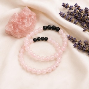 Puede incluir: Dos pulseras de cuentas con cuentas redondas de color rosa pálido y una pequeña sección de cuatro cuentas de lava negra texturizadas. Las pulseras están dispuestas en un patrón circular, mostrando el contraste entre la piedra clara y la roca volcánica oscura.