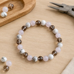 Puede incluir: Una pulsera de cuentas con un patr&oacute;n repetitivo de cuentas redondas de m&aacute;rmol blanco, cuentas de color rosa claro transl&uacute;cido y cuentas de cuarzo ahumado marr&oacute;n oscuro. La pulsera est&aacute; dispuesta en c&iacute;rculo con cuentas sueltas cerca.