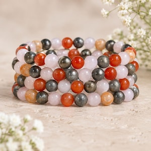 May include: A multi-strand bracelet composed of round beads in alternating colors of translucent pale pink, opaque bright orange-red, and metallic dark grey. The beads are arranged in a repeating pattern across several parallel elasticized strands.