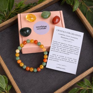 May include: A pink cardboard box labeled soothing crystals containing four loose polished stones in yellow, green, dark green, and red. A beaded bracelet with alternating orange, yellow, and green beads sits beside a white card titled Crystals for Creativity.