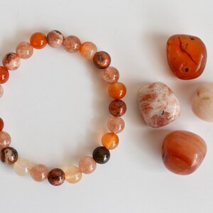 May include: A bracelet made of orange, brown, and white beads. There are four loose stones next to the bracelet: two orange, one white, and one brown.