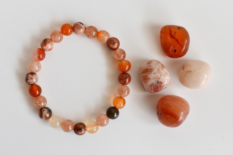 May include: A bracelet made of orange, brown, and white beads. There are four loose stones next to the bracelet: two orange, one white, and one brown.