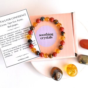 Pode incluir: Um bracelete feito com pedras de olho de tigre, jaspe vermelho e citrino. O bracelete est&aacute; em uma pequena caixa com um cart&atilde;o que diz "Cristais para a confian&ccedil;a" e lista as pedras usadas no bracelete. H&aacute; tamb&eacute;m quatro pedras soltas: olho de tigre, jaspe vermelho, citrino e pirita.