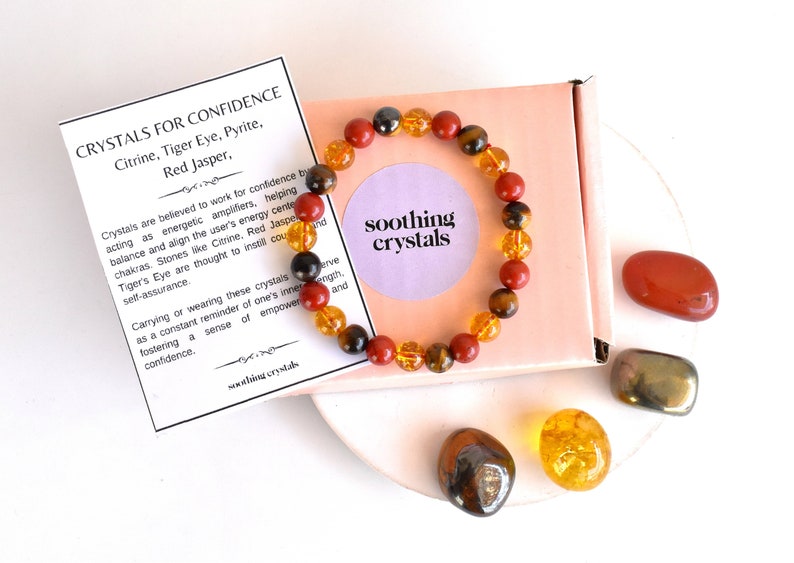 Pode incluir: Um bracelete feito com pedras de olho de tigre, jaspe vermelho e citrino. O bracelete est&aacute; em uma pequena caixa com um cart&atilde;o que diz "Cristais para a confian&ccedil;a" e lista as pedras usadas no bracelete. H&aacute; tamb&eacute;m quatro pedras soltas: olho de tigre, jaspe vermelho, citrino e pirita.