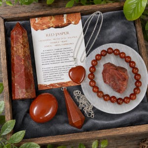 May include: A collection of red jasper items including a tall obelisk, a smooth palm stone, a heart-shaped pendant on a silver chain, a faceted pendulum on a silver chain, a beaded bracelet, and a raw stone piece, accompanied by an informational card.