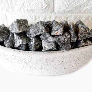 May include: A white ceramic bowl filled with gray and black stones. The bowl has a speckled texture and a rounded shape.