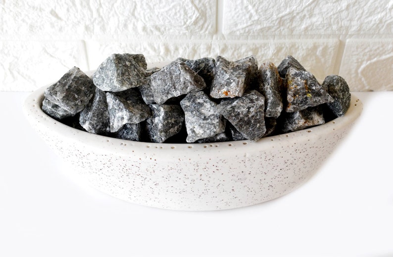 May include: A white ceramic bowl filled with gray and black stones. The bowl has a speckled texture and a rounded shape.