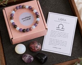 Pode incluir: Uma pulseira de contas roxas, castanhas e brancas translúcidas sobre uma caixa rosa com a etiqueta soothing crystals. Ao lado, um cartão branco com o texto LIBRA e quatro pedras soltas: uma branca, uma rosa manchada, uma roxa e uma preta.
