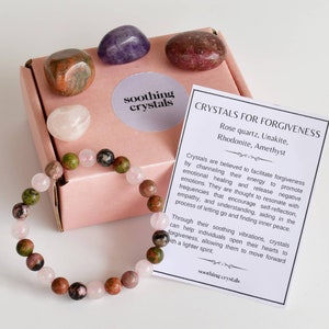 May include: A pink box with a variety of crystals inside, including rose quartz, amethyst, rhodonite, and unakite. The box also includes a bracelet made of the same crystals. The text on the card reads "Crystals for Forgiveness" and lists the types of crystals included. The text also describes the healing properties of the crystals.