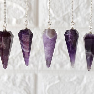 Könnte beinhalten: Sechs Amethyst-Kristallpendel mit silbernen Ketten. Die Pendel sind alle spitz zulaufend und haben einen violetten Farbton.