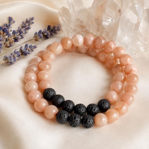 May include: A double-strand beaded bracelet featuring smooth, round, peach-colored stone beads and a segment of textured, porous black lava beads. The bracelet is arranged in a circular shape, showcasing the contrast between the light and dark beads.