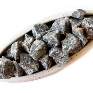 May include: A white ceramic oval dish filled with gray and black stones. The stones are smooth and have a natural, rough texture.