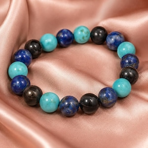 May include: A beaded bracelet featuring a repeating pattern of round, polished stones in shades of turquoise blue, deep royal blue, and glossy black. The beads are strung together in a circular arrangement, creating a continuous, uniform band.