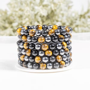 Puede incluir: Una pila de seis pulseras de cuentas con cuentas redondas alternas de piedra negra pulida y ojo de tigre marrón dorado. Las pulseras están dispuestas en una columna vertical, mostrando un patrón repetitivo de tonos oscuros y terrosos.