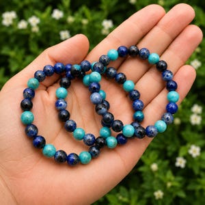 May include: Two stretch bracelets composed of round beads in varying shades of deep navy blue, royal blue, and bright turquoise. The beads feature natural mottled patterns and smooth, polished surfaces, arranged in a repeating color sequence.