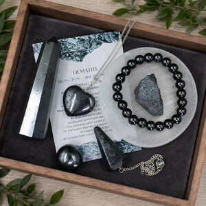 May include: A collection of hematite items including a polished tower, a heart-shaped pendant on a silver chain, a round sphere, a beaded bracelet, and a raw stone fragment. A white information card titled Hematite is included in the set.