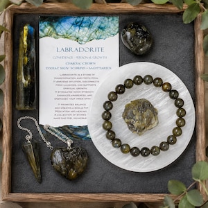 May include: A collection of labradorite items including a polished tower, a heart-shaped pendant on a silver chain, a pendulum, a raw stone, and a beaded bracelet on a white circular plate, accompanied by an informational card about labradorite.