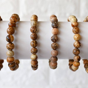 May include: Six brown and beige gemstone bracelets with round beads. The bracelets are displayed on a white surface.