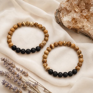 Op de afbeelding: Twee elastische armbanden met ronde kralen. Elke armband bestaat uit bruin en beige gemarmerde jaspis kralen gecombineerd met een sectie van vijf poreuze zwarte lavakralen. De kralen zijn in een cirkel op een lichte stof geplaatst.