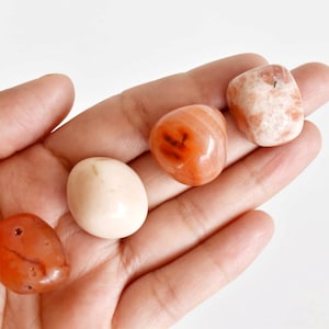 May include: Four tumbled stones in a hand. The stones are various shades of orange, pink, and white.