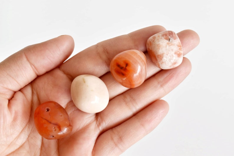 May include: Four tumbled stones in a hand. The stones are various shades of orange, pink, and white.