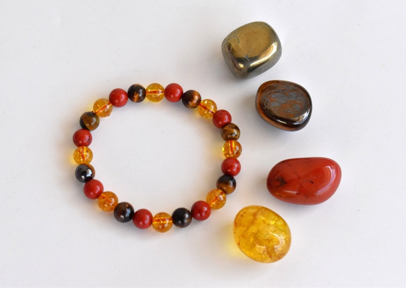 Pode incluir: Um bracelete feito com contas vermelhas, amarelas e marrons. O bracelete &eacute; rodeado por pedras soltas das mesmas cores.
