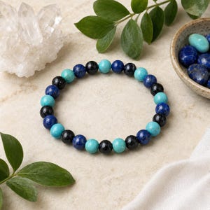 May include: A beaded bracelet featuring a repeating pattern of round polished beads in shades of turquoise blue, deep royal blue, and glossy black. The beads are strung on an elastic cord, creating a circular shape with a smooth, uniform texture.