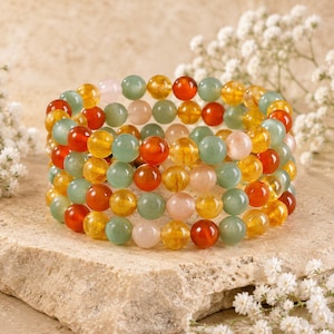 May include: A multi-strand beaded bracelet featuring polished spherical beads in shades of translucent green, opaque orange, deep red, and pale pink. The beads are arranged in a circular formation, creating a layered appearance with a smooth texture.