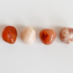 May include: Four tumbled stones in a row on a white background. The stones are a light pink, a dark orange, a light orange, and a white with brown streaks.