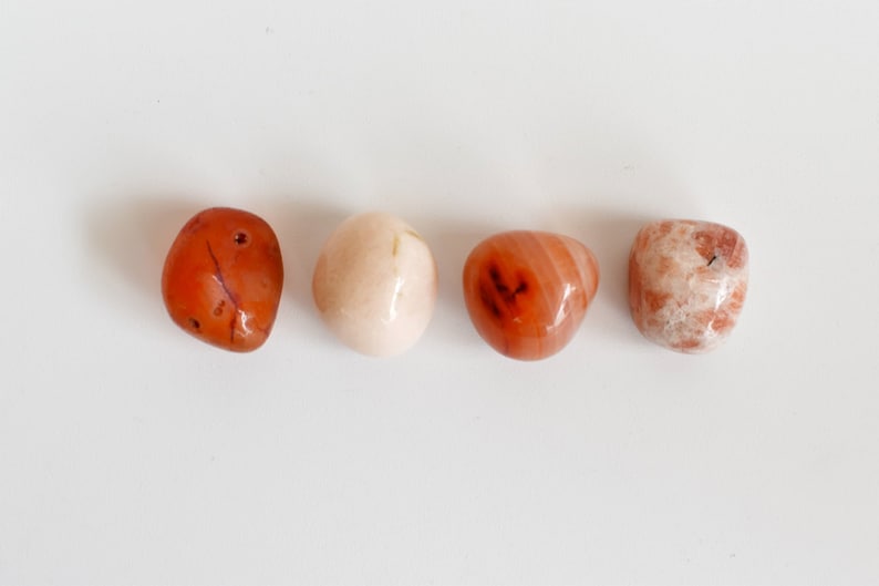 May include: Four tumbled stones in a row on a white background. The stones are a light pink, a dark orange, a light orange, and a white with brown streaks.
