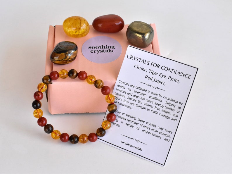 Pode incluir: Um conjunto de cristais para a confian&ccedil;a, incluindo citrino, olho de tigre, pirita e jaspe vermelho. Os cristais est&atilde;o dispostos numa caixa rosa com uma pulseira feita das mesmas pedras. O cart&atilde;o diz "Cristais para a confian&ccedil;a" e lista as pedras inclu&iacute;das no conjunto.