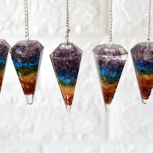May include: Five rainbow chakra crystal pendulums with silver chains. Each pendulum is made of clear resin with layers of different colored crystals, representing the seven chakras.