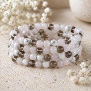 Puede incluir: Una pulsera de cuentas de varias hebras con un patr&oacute;n repetitivo de cuentas redondas y lisas de cuarzo ahumado transl&uacute;cido, cuarzo rosa opaco y howlita blanca veteada, dispuestas en un dise&ntilde;o circular y el&aacute;stico.