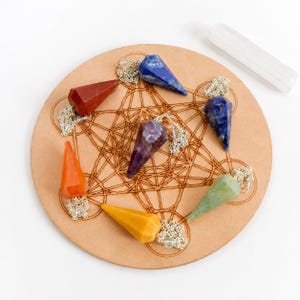 May include: Seven faceted gemstone pendulums in red, orange, yellow, green, purple, and two shades of blue, each attached to a silver-toned chain. The pendulums are arranged on a circular wooden board engraved with a geometric star pattern.