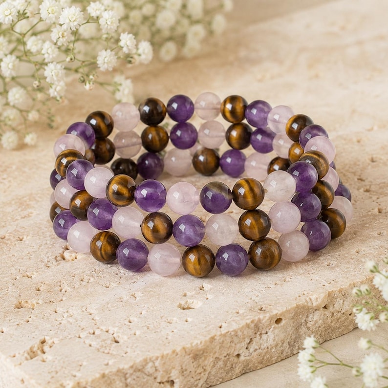 May include: A triple-strand beaded bracelet featuring alternating round polished beads in shades of purple amethyst, brown tiger's eye, and pale pink rose quartz. The beads are strung on an elastic cord, creating a layered circular design.